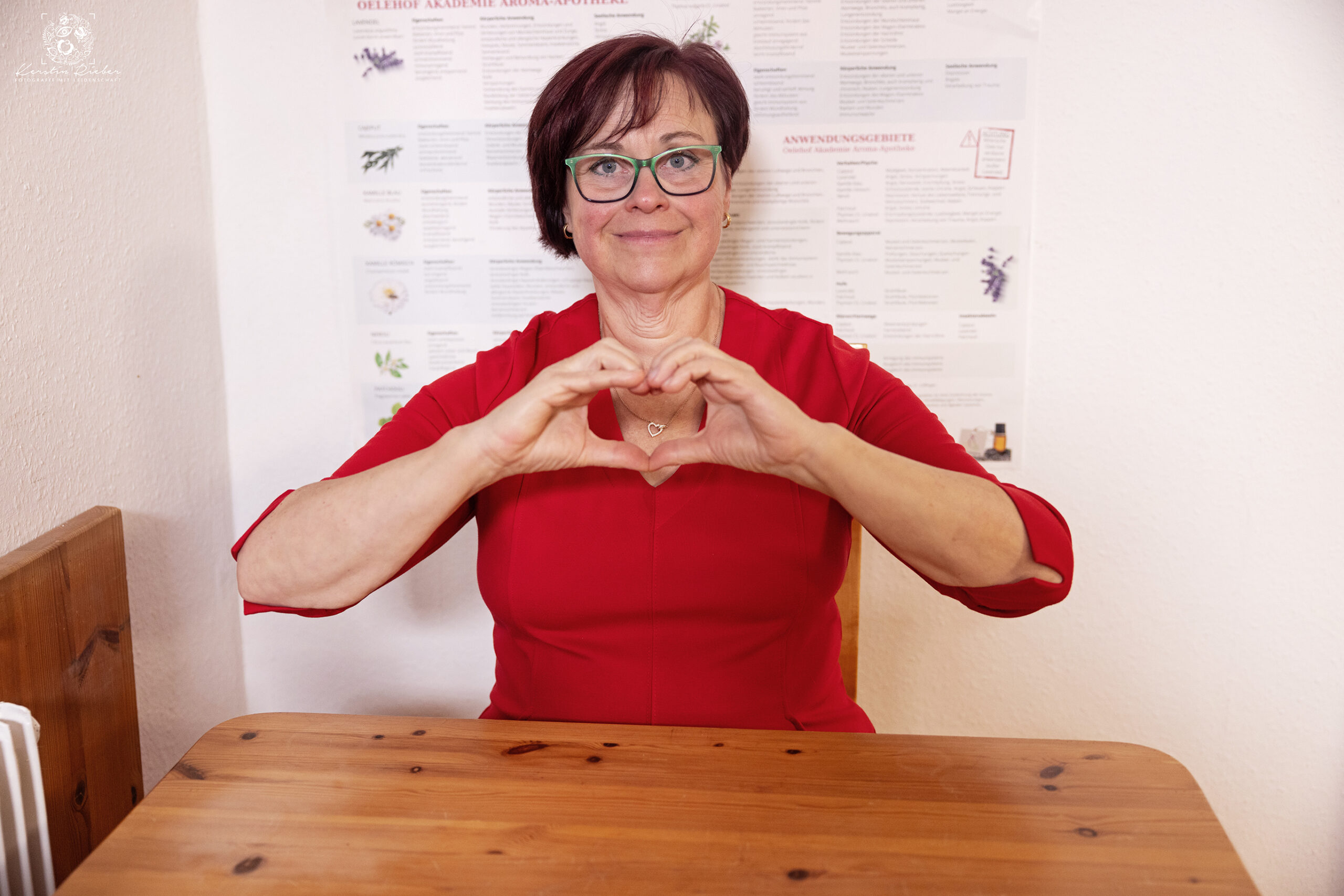 Monika sitzt im roten Kleid am Tisch und formt mit den Händen ein Herz